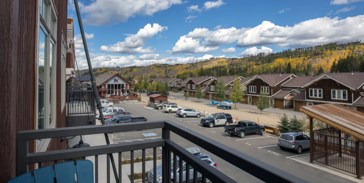A private balcony with a railing overlooking a tranquil mountain landscape. Residential buildings are visible in the background, and the space is perfect for relaxing, socializing, or grilling while enjoying the alpine scenery.