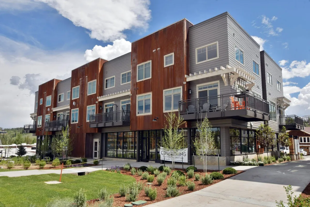 The image shows a modern exterior facade with clean architectural lines, expansive windows, lush green landscaping, and inviting outdoor seating areas. The scene reflects a lively yet welcoming community atmosphere, with hints of social gatherings and neighbors relaxing outdoors. The overall vibe is upbeat and connected, suggestive of convenient, community-centric living in a mountain town.
