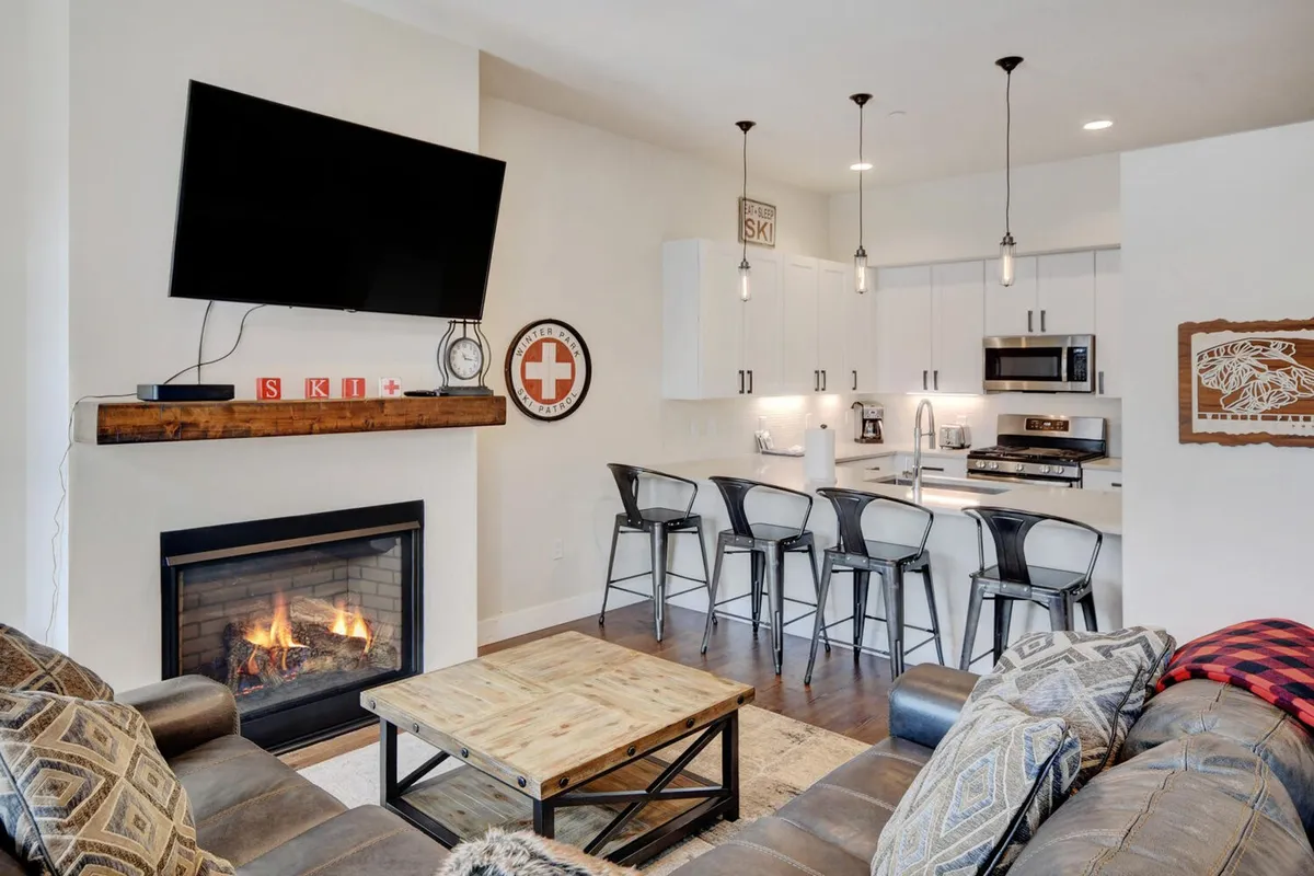The hero image shows a stylish, open-concept living space with a crackling fireplace, a spacious kitchen island, a plush sectional sofa, and a large TV. The room exudes warmth and comfort, inviting guests to relax with hot cocoa, cook together, and enjoy cozy movie nights after adventuring in the Colorado mountains.