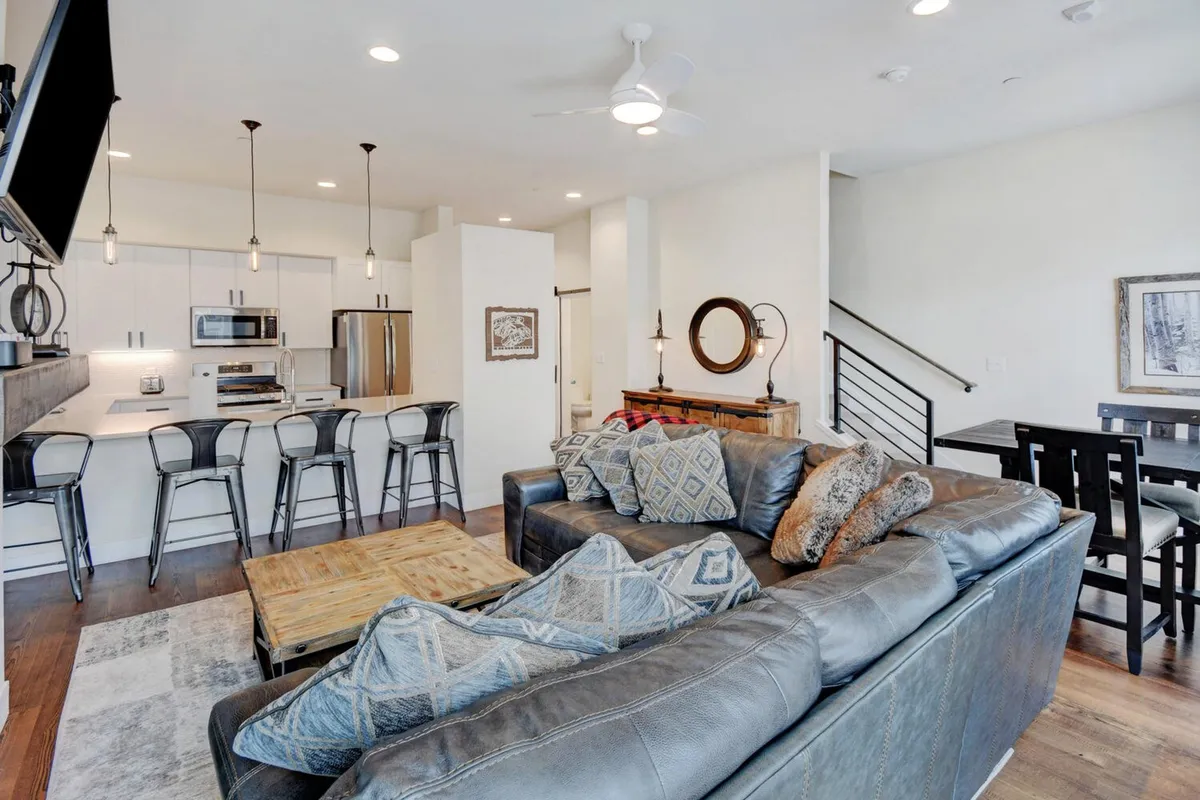 This image presents an open, welcoming interior featuring a plush leather sofa, wooden coffee table, bar stools at the kitchen counter, and a large dining table. The scene suggests an atmosphere ideal for socializing and relaxing, with plenty of natural light and thoughtful design elements that emphasize comfort and togetherness.