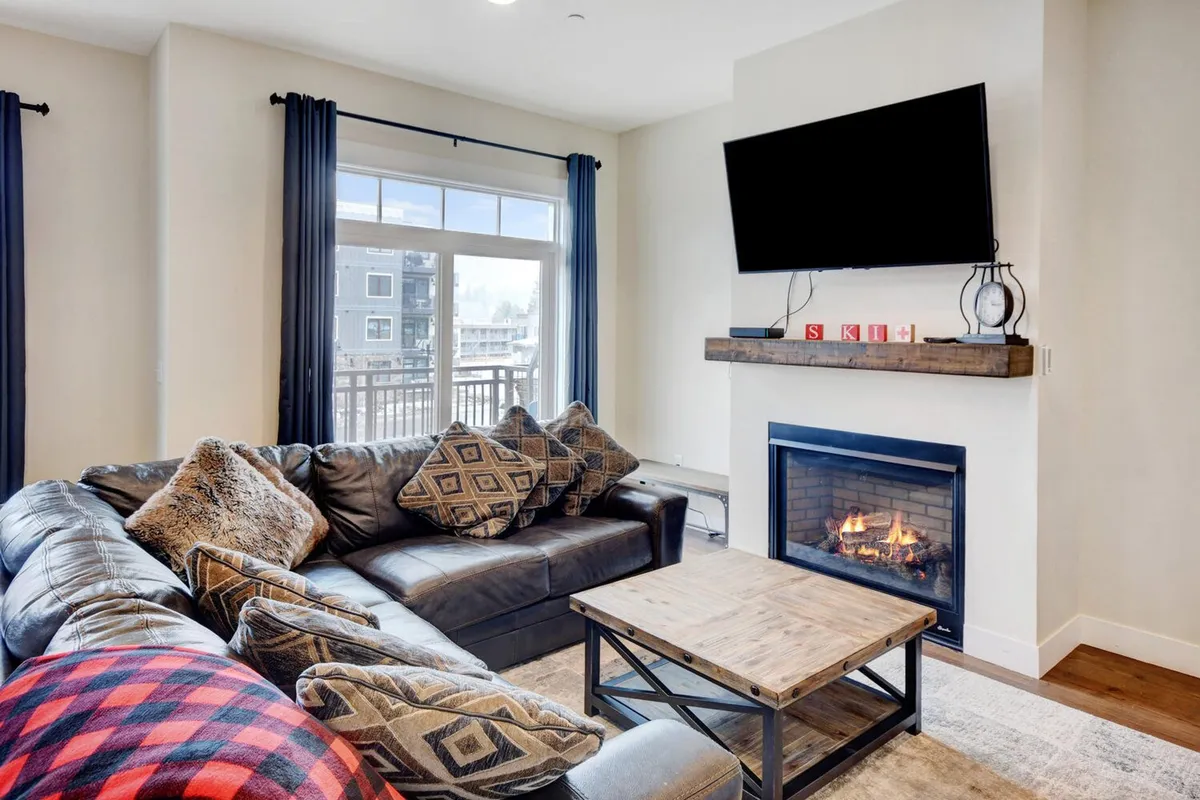A cozy corner featuring a leather sofa, a crackling fireplace, a wooden coffee table, and a large window that provides scenic outdoor views. The setting is perfect for relaxing after mountain adventures, with the option to watch TV, read, or simply enjoy the warmth and ambiance of the fire.