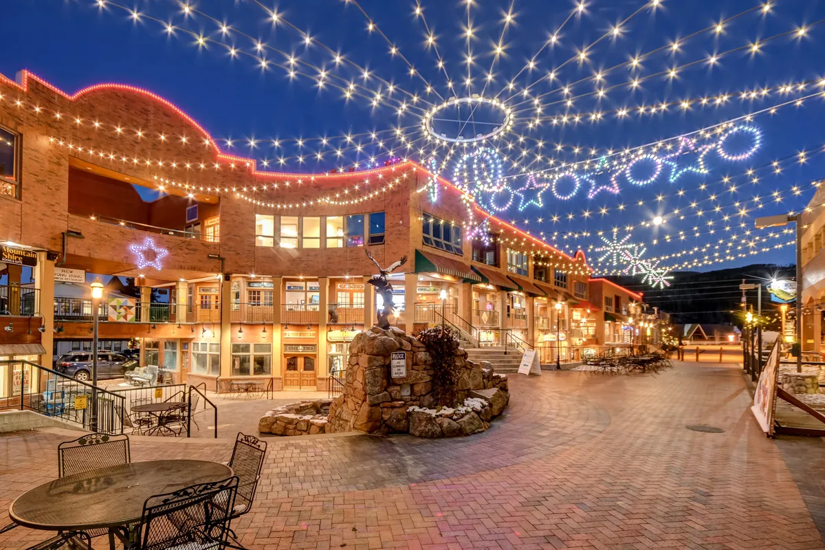 A lively exterior evening scene with twinkling string lights, decorative stars, a stone fountain, and adjacent storefronts, evoking a sense of local community gatherings and festive outdoor fun.