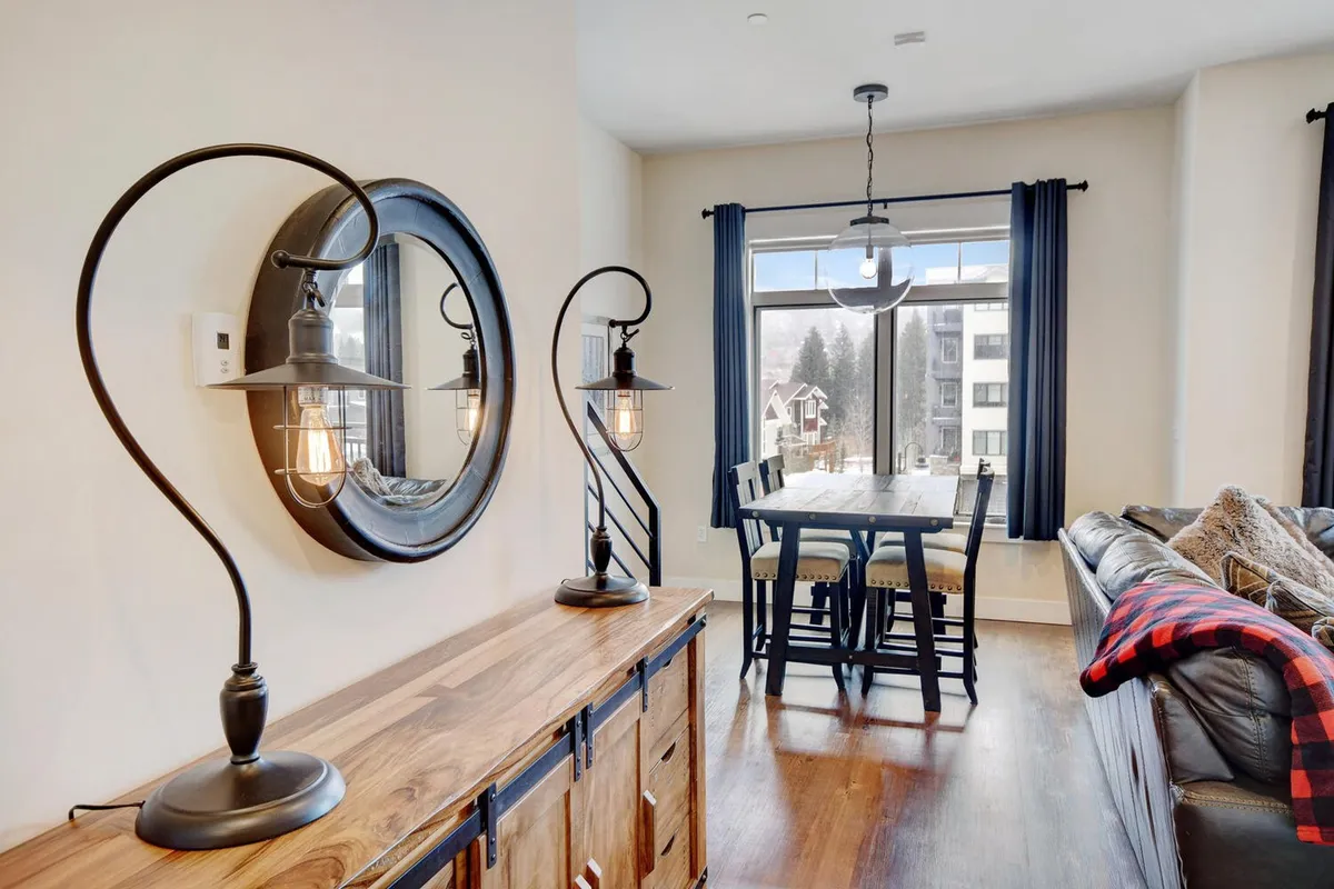 A cozy dining area anchored by a rustic wooden sideboard, a large round mirror, and a spacious dining table surrounded by chairs. Overhead, a pendant light fixture casts a warm glow, while a comfortable sofa with a throw blanket in the background hints at a relaxed, communal setting.