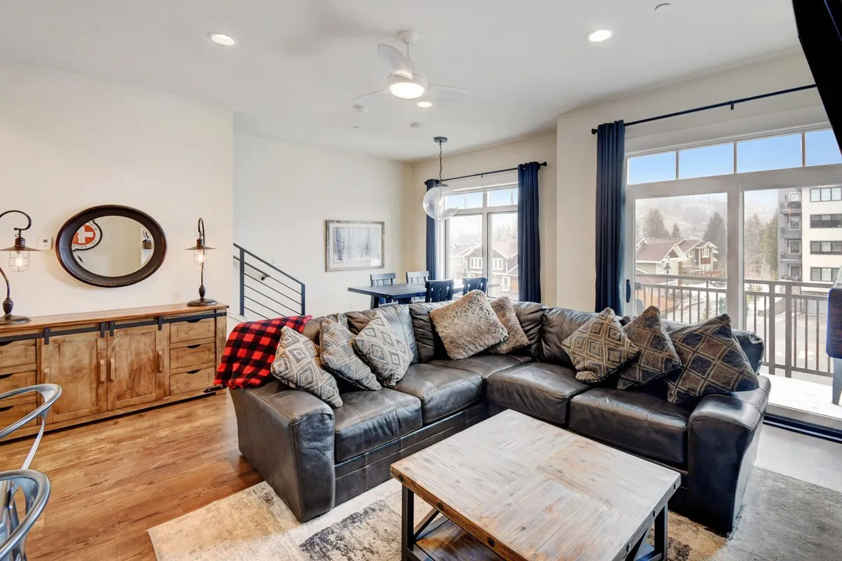 A spacious living area featuring a large sectional sofa with decorative pillows, a wooden coffee table, and expansive windows that fill the space with natural light. The windows offer a glimpse of the scenic outdoors, and the setup is perfect for relaxing, entertaining guests, or enjoying a cozy movie night. The room exudes a warm and welcoming atmosphere, ideal for family gatherings or unwinding after a day of adventure.