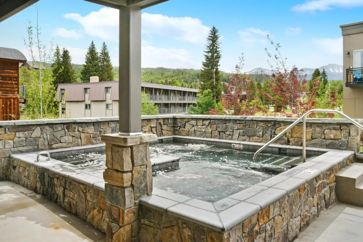 A spacious outdoor hot tub surrounded by natural stone walls and mature trees, with mountains visible in the background. The tranquil setting shows guests relaxing in the steaming water, with soft evening light casting a warm glow over the scene.