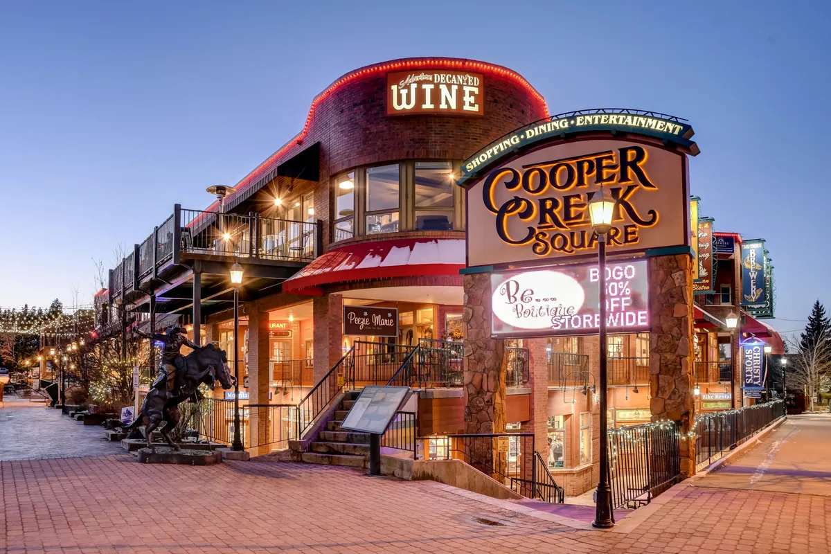 The photo captures the entrance to Cooper Creek Square, highlighted by decorative string lights and a welcoming staircase. The area is bustling with boutique shops, a wine shop entrance, and evening ambiance, inviting exploration, dining, and shopping experiences. The festive vibe reflects the dynamic energy of downtown Winter Park.