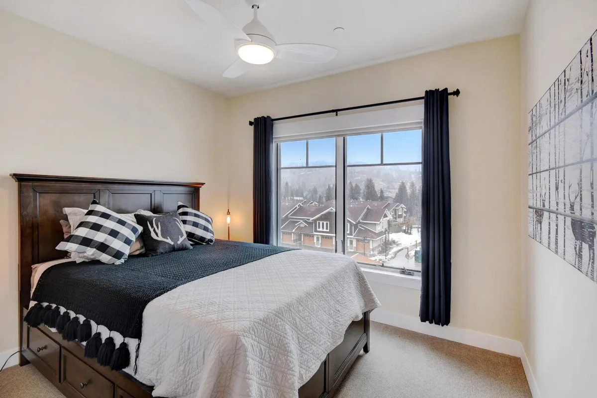 A serene bedroom featuring a comfortable bed positioned near a large window, with decorative artwork enhancing the peaceful ambiance. The natural light and scenic view through the window suggest the opportunity to wake up to beautiful mountain vistas.